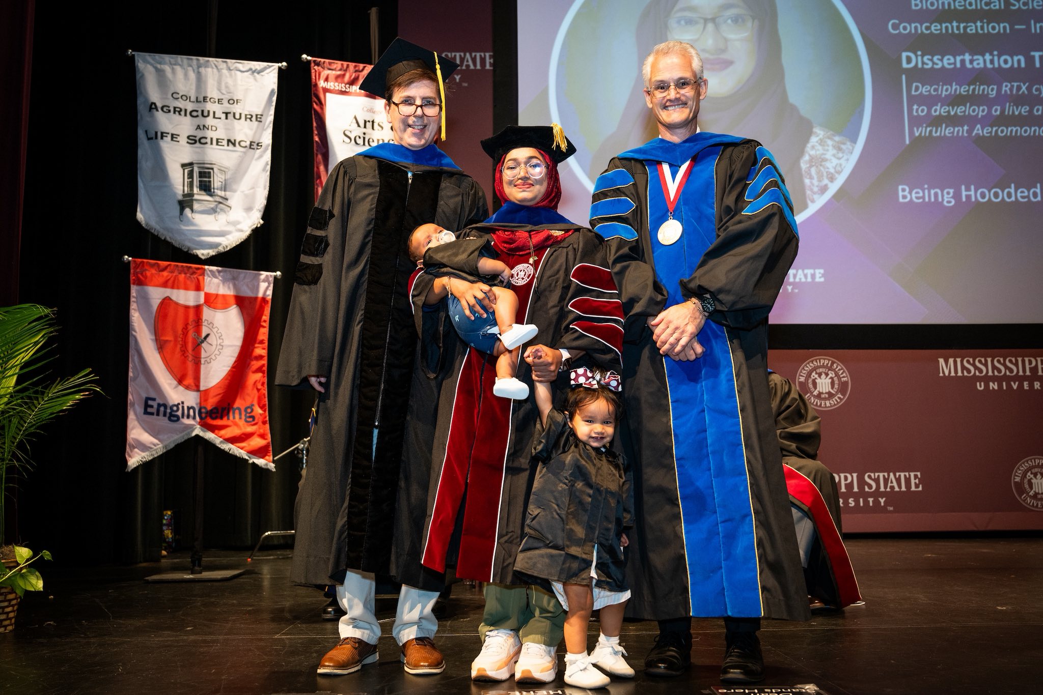 A woman graduating with two children surrounded by professors.