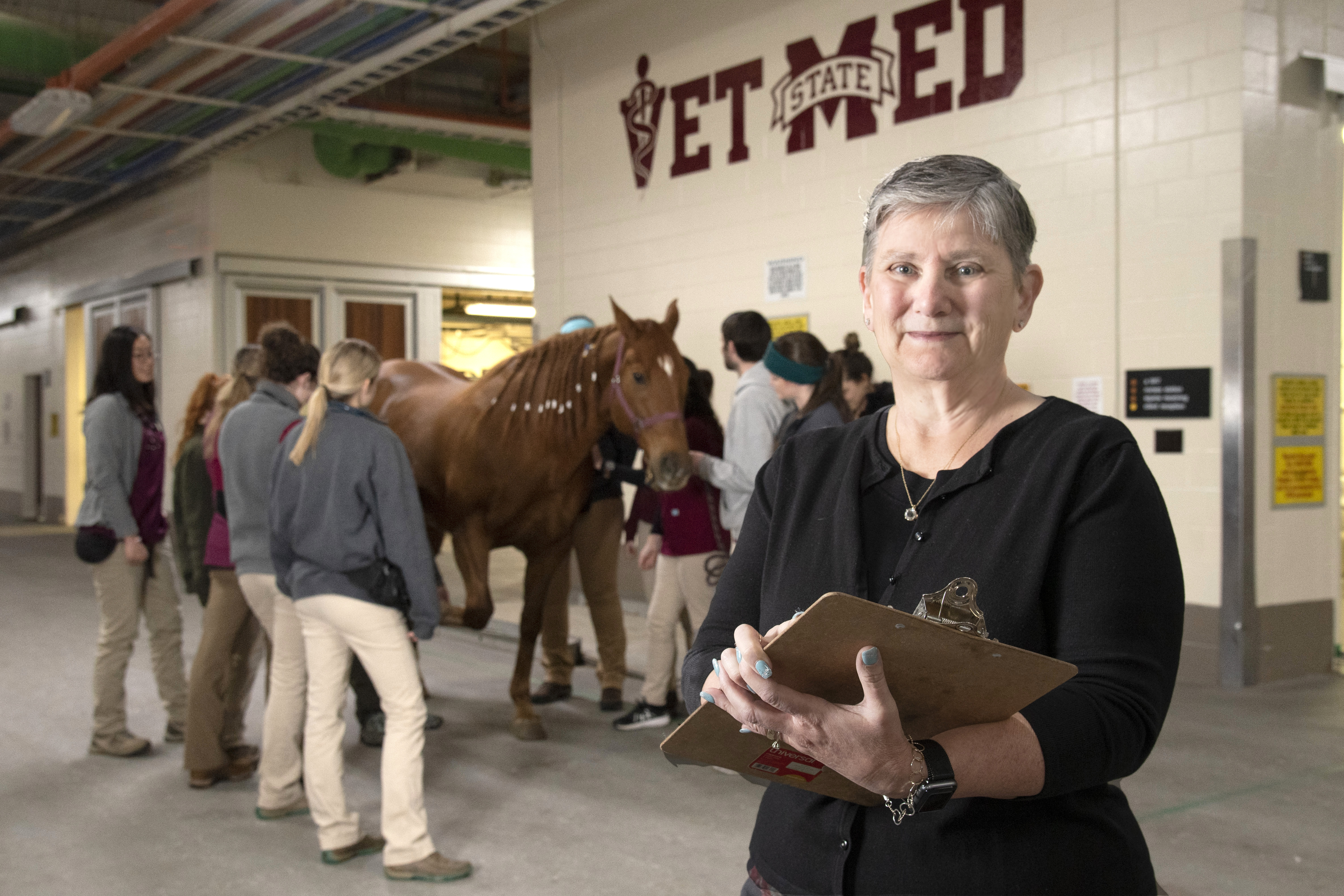 A woman poses with a clipboard in front of a horse and several students