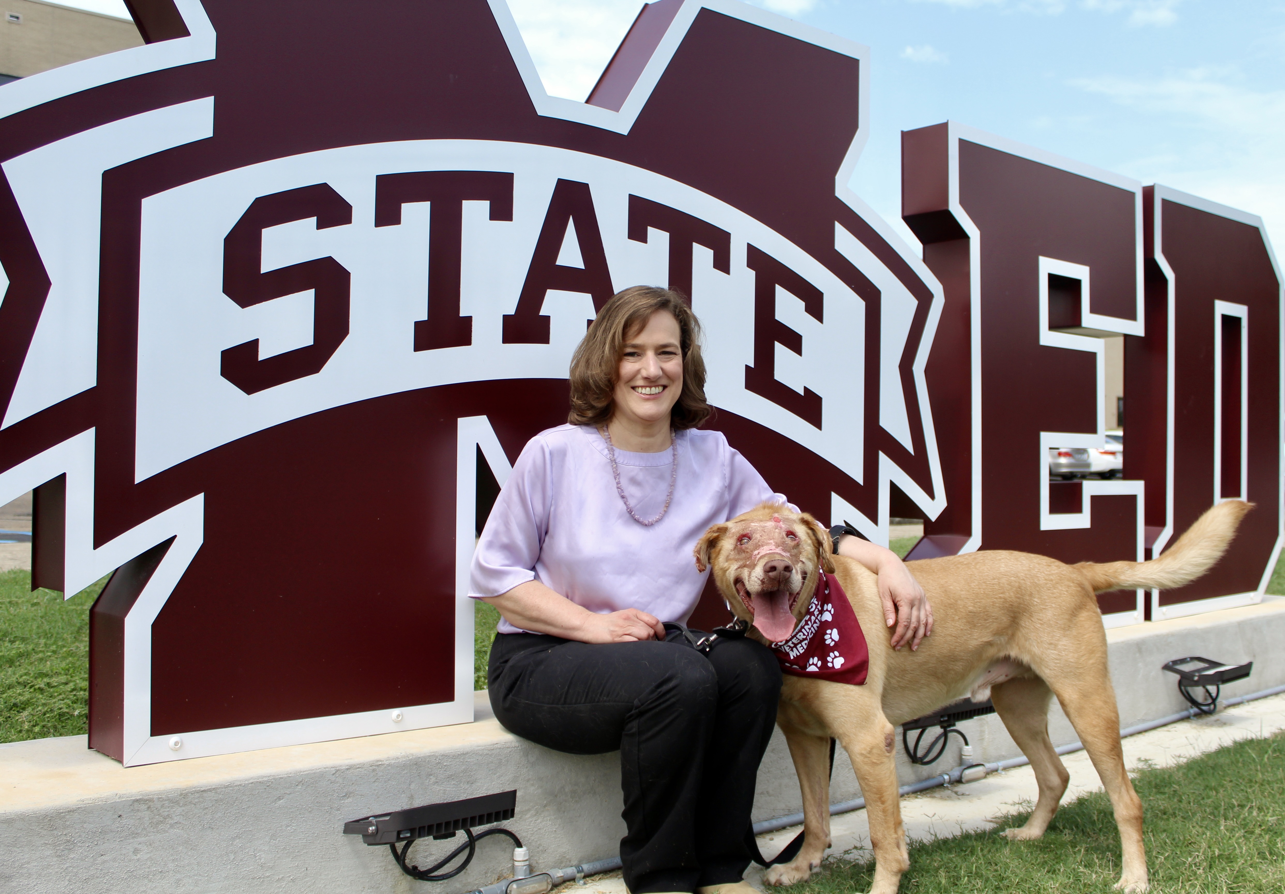 Buddy the Miracle Lab and his owner, Dr. Betsy Swanson, sit together in front of the VetMed sign located at the front of the Mississippi State University College of Veterinary Medicine Wise Center.
